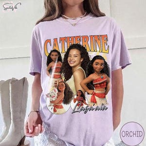 K&ouml;nnte beinhalten: Ein hellviolettes Kurzarm-T-Shirt mit einem Grafikdruck des Namens Catherine in fetten orangefarbenen Buchstaben &uuml;ber einer Collage aus vier animierten Figuren und einem zentralen Portr&auml;t einer Person mit langem, dunklem, lockigem Haar.