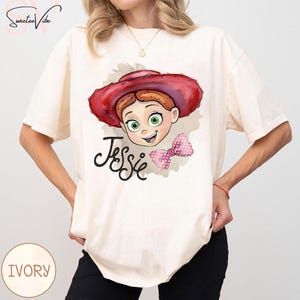 Puede incluir: Camiseta de color marfil con una ilustraci&oacute;n de dibujos animados de Jessie de Toy Story. El dise&ntilde;o incluye la cara de Jessie, un sombrero rojo, un lazo rosa y el nombre "Jessie" en cursiva. La camiseta es de corte holgado.
