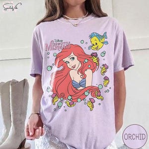 May include: A light purple t-shirt featuring a graphic of Ariel from Disney's The Little Mermaid. Ariel has red hair and a blue seashell bra. The shirt also features Flounder and seahorse illustrations.