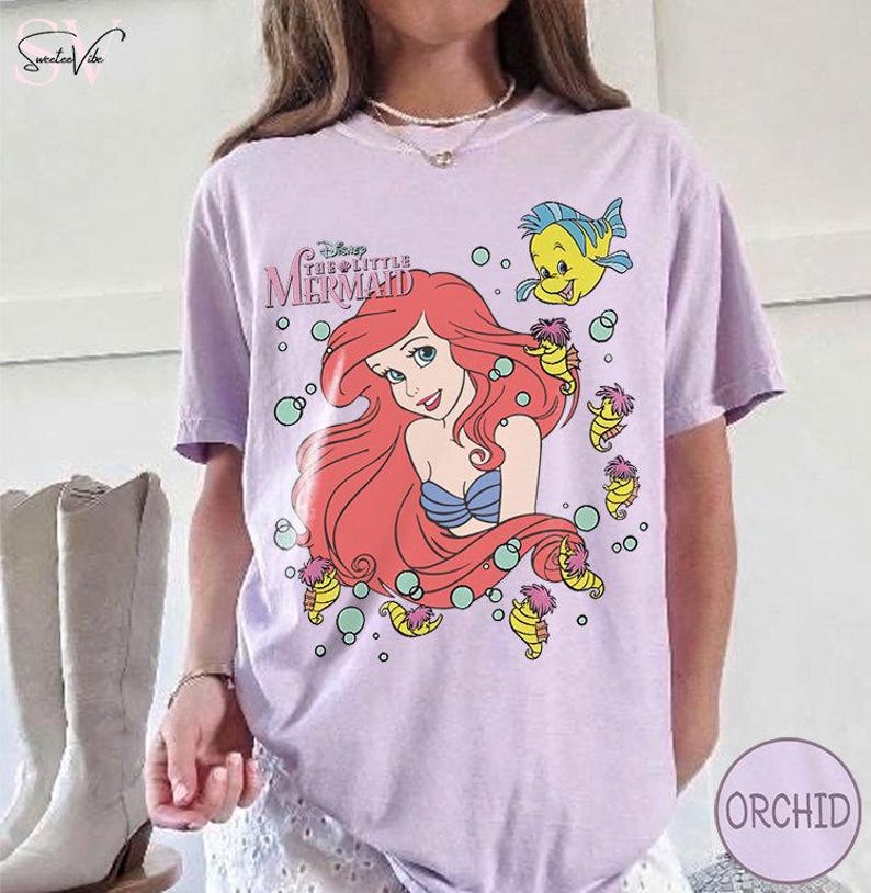 May include: A light purple t-shirt featuring a graphic of Ariel from Disney's The Little Mermaid. Ariel has red hair and a blue seashell bra. The shirt also features Flounder and seahorse illustrations.