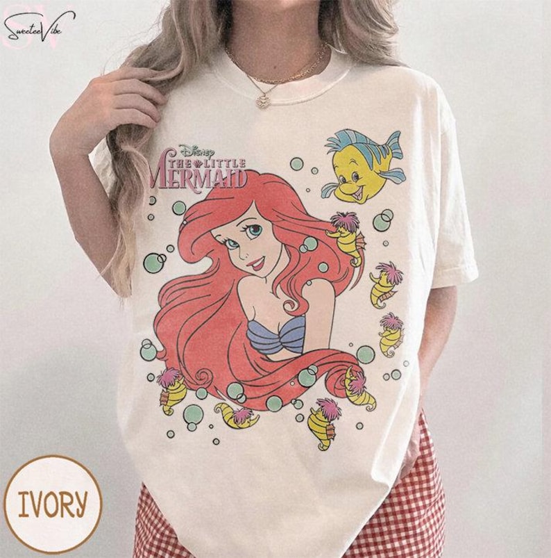 May include: Ivory-colored t-shirt featuring a graphic of Ariel from Disney's The Little Mermaid. The design includes Ariel with red hair, a blue top, and a yellow fish, surrounded by seahorses and bubbles. The text "Disney The Little Mermaid" is above Ariel.