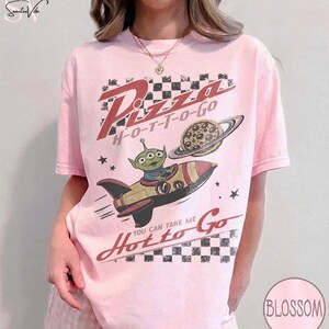 May include: Pink t-shirt with a graphic featuring an alien in a rocket ship, a pizza planet, and the words "Pizza H-O-T-T-O-Go" and "You can take me Hot to Go". The design has a retro, cartoonish style.
