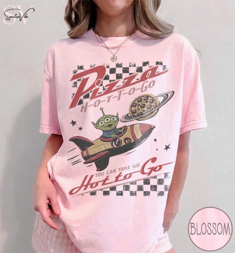 May include: Pink t-shirt with a graphic featuring an alien in a rocket ship, a pizza planet, and the words "Pizza H-O-T-T-O-Go" and "You can take me Hot to Go". The design has a retro, cartoonish style.