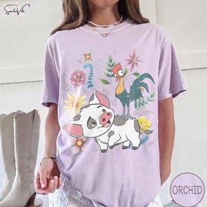 Puede incluir: Camiseta lila claro con un dise&ntilde;o de cerdo y gallo de dibujos animados, rodeado de flores. El cerdo es blanco con manchas grises y orejas rosas, y el gallo es verde, amarillo y rojo. La palabra "ORCHID" est&aacute; en un c&iacute;rculo en la parte inferior derecha.