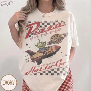 May include: Ivory-colored t-shirt with a retro graphic design. The design features the words "Pizza H-O-T-T-O-Go" in red, a cartoon alien in a rocket ship, and a pizza planet. The text "You Can Take Me Hot to Go" is also included.