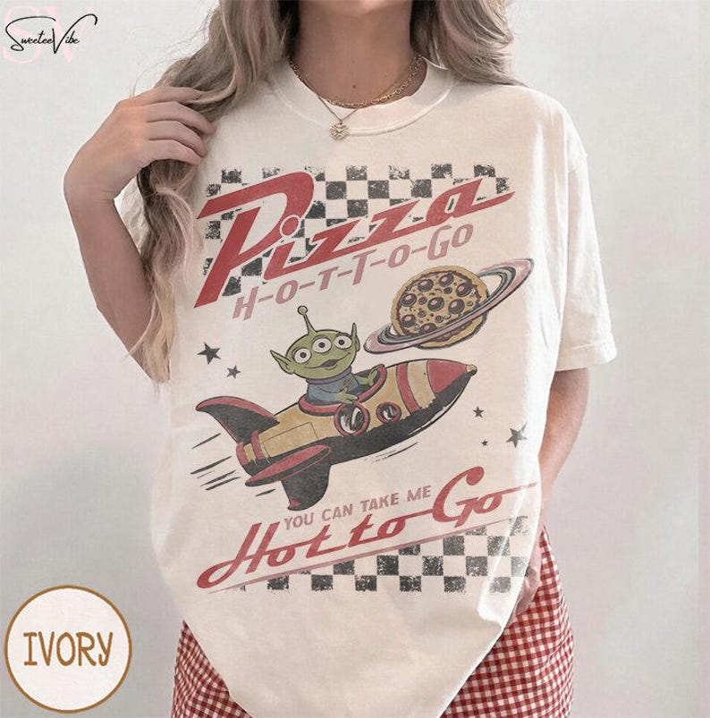 May include: Ivory-colored t-shirt with a retro graphic design. The design features the words "Pizza H-O-T-T-O-Go" in red, a cartoon alien in a rocket ship, and a pizza planet. The text "You Can Take Me Hot to Go" is also included.