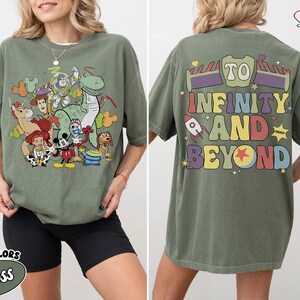 Op de afbeelding: Olijfgroen T-shirt met een kleurrijke afbeelding van Toy Story-personages op de voorkant. De achterkant van het shirt toont de zin "To Infinity and Beyond" in een retro lettertype. Het shirt is een comfort kleur.