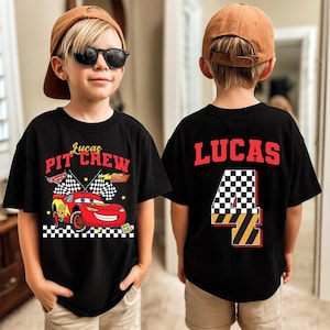 May include: Black t-shirt with a red race car graphic and the text "Lucas Pit Crew". The back of the shirt has the name "LUCAS" and the number "4" in a checkered flag design. The shirt is worn by a child.