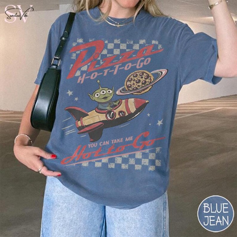 May include: Blue t-shirt with a retro graphic featuring an alien in a rocket ship, a pizza planet, and the text "Pizza H-O-T-T-O-Go". The design includes stars and a checkered pattern, creating a vintage aesthetic.