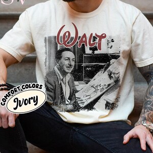 May include: Ivory-colored t-shirt featuring a black and white image of Walt Disney holding a drawing. The word "Walt" is printed in burgundy script above the image. The shirt also has the words "Comfort Colors" and "Ivory" printed on it.