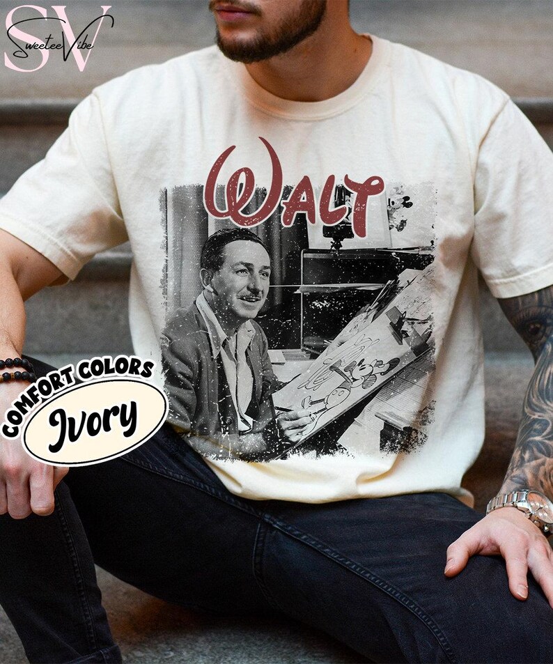 May include: Ivory-colored t-shirt featuring a black and white image of Walt Disney holding a drawing. The word "Walt" is printed in burgundy script above the image. The shirt also has the words "Comfort Colors" and "Ivory" printed on it.