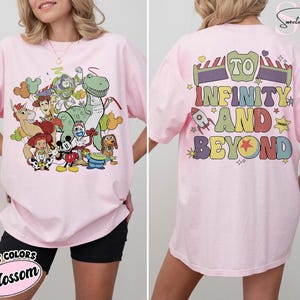Op de afbeelding: Roze T-shirt met een kleurrijke afbeelding van Toy Story-personages op de voorkant. De achterkant van het shirt heeft de tekst "To Infinity and Beyond". Het shirt is een Comfort Colors Blossom.