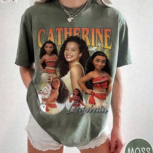 K&ouml;nnte beinhalten: Ein moosgr&uuml;nes Kurzarm-T-Shirt mit einem Grafikdruck der Figur Moana in verschiedenen Posen, unter anderem mit einem Schwein. Der Text CATHERINE ist oben in gro&szlig;en orangefarbenen Buchstaben gedruckt, und der Name Moana steht in Schreibschrift.
