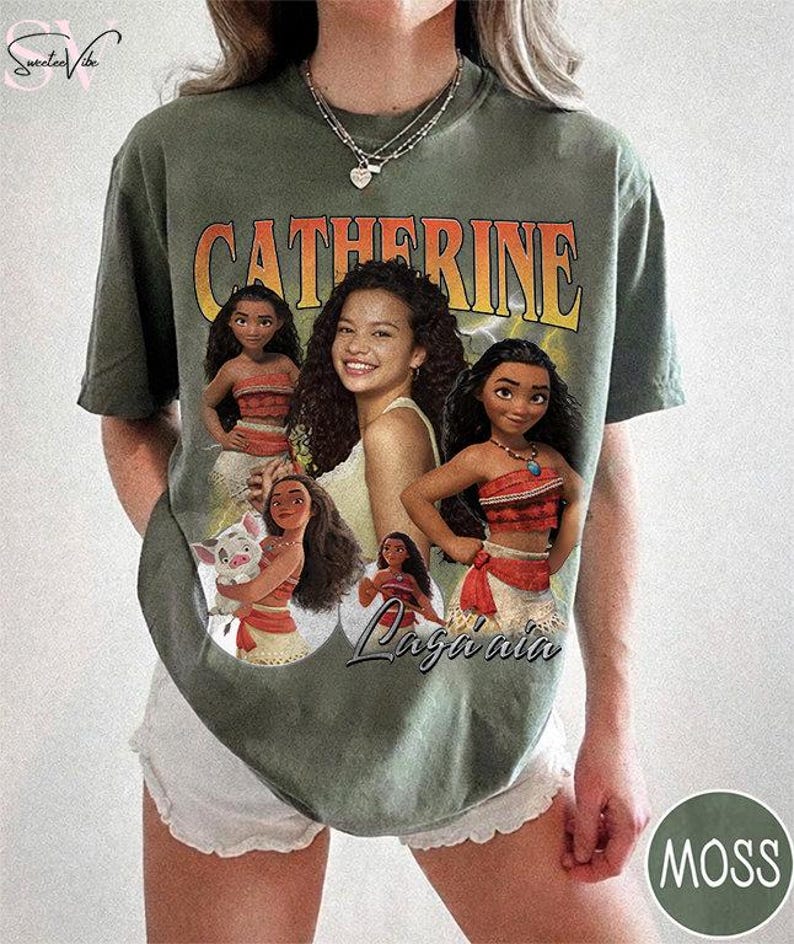 K&ouml;nnte beinhalten: Ein moosgr&uuml;nes Kurzarm-T-Shirt mit einem Grafikdruck der Figur Moana in verschiedenen Posen, unter anderem mit einem Schwein. Der Text CATHERINE ist oben in gro&szlig;en orangefarbenen Buchstaben gedruckt, und der Name Moana steht in Schreibschrift.
