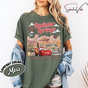 Puede incluir: Una camiseta verde musgo con un gr&aacute;fico de Radiator Springs y Rayo McQueen. El dise&ntilde;o incluye el texto "Radiator Springs" en rojo, junto con im&aacute;genes de la ciudad y el personaje. La camiseta est&aacute; etiquetada como "Comfort Colors Moss".