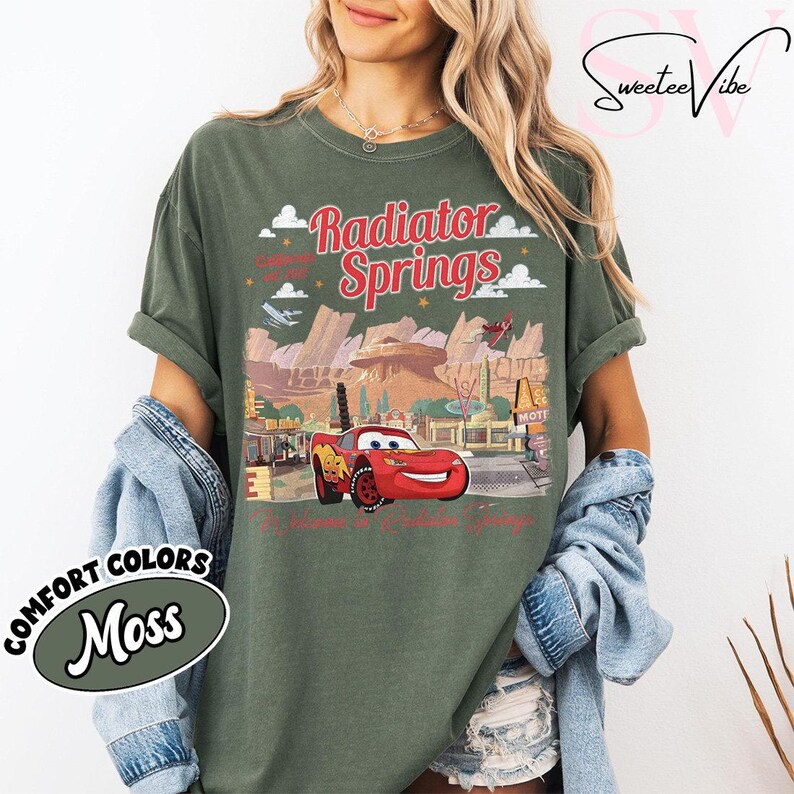 Puede incluir: Una camiseta verde musgo con un gr&aacute;fico de Radiator Springs y Rayo McQueen. El dise&ntilde;o incluye el texto "Radiator Springs" en rojo, junto con im&aacute;genes de la ciudad y el personaje. La camiseta est&aacute; etiquetada como "Comfort Colors Moss".