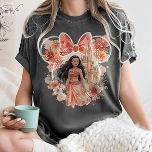 May include: Dark gray t-shirt featuring a graphic of a cartoon character in a dress, with a castle and floral design. The design is set within a Mickey Mouse ear outline with a bow. The shirt has short sleeves.