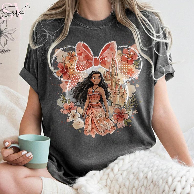 May include: Dark gray t-shirt featuring a graphic of a cartoon character in a dress, with a castle and floral design. The design is set within a Mickey Mouse ear outline with a bow. The shirt has short sleeves.