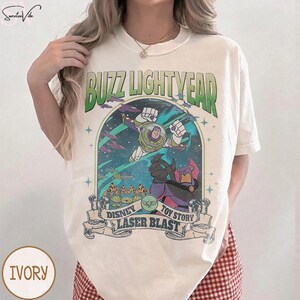 Puede incluir: Camiseta de color marfil con un gr&aacute;fico de Buzz Lightyear de Toy Story. El dise&ntilde;o incluye el texto "Buzz Lightyear" en verde y "Disney Toy Story Laser Blast" en una pancarta. La camiseta tiene cuello redondo y mangas cortas.