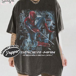 May include: A charcoal gray short-sleeve t-shirt featuring a graphic print of Spider-Man in a dynamic pose against a city background. The text "SPIDER-MAN A BRAND NEW DAY" is printed in white below the character. The shirt has a crew neckline.
