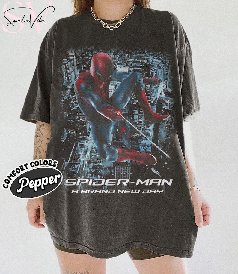 May include: A charcoal gray short-sleeve t-shirt featuring a graphic print of Spider-Man in a dynamic pose against a city background. The text "SPIDER-MAN A BRAND NEW DAY" is printed in white below the character. The shirt has a crew neckline.