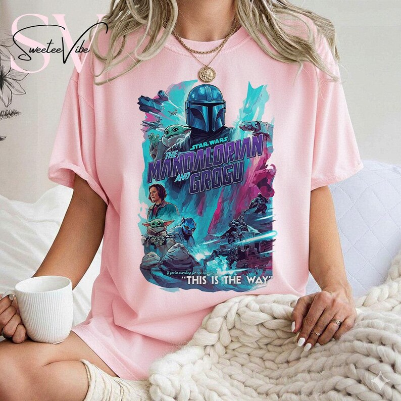 May include: Pink t-shirt featuring a graphic from the Star Wars series, "The Mandalorian and Grogu." The design includes characters and the phrase "THIS IS THE WAY." The shirt has short sleeves and a crew neck.