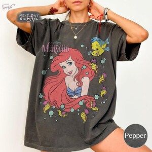 May include: Dark gray t-shirt featuring a graphic of Ariel from Disney's The Little Mermaid. The design includes Ariel with red hair, a blue seashell bra, and the text "The Little Mermaid". Additional characters include Flounder and seahorses.