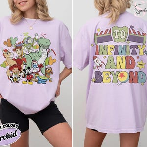 Op de afbeelding: Een lichtpaars T-shirt met een kleurrijke afbeelding van Toy Story-personages op de voorkant. De achterkant van het shirt heeft de tekst "To Infinity and Beyond" in een regenboogontwerp. Het shirt is een Comfort Colors Orchid.