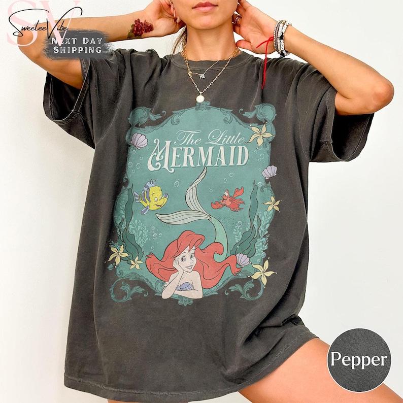 May include: Dark gray t-shirt featuring a graphic from "The Little Mermaid." The design includes Ariel, Flounder, and Sebastian, with the title in a teal frame. The shirt is a relaxed fit.