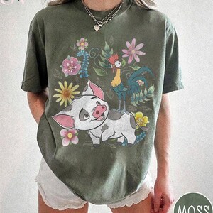 Puede incluir: Camiseta verde musgo con un dise&ntilde;o de cerdo de dibujos animados, gallo y flores. El cerdo es blanco con manchas grises y orejas rosas. El gallo es amarillo y azul. Las flores son rosas, amarillas y azules. La camiseta tiene cuello redondo y mangas cortas.