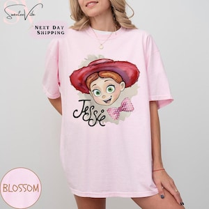 Puede incluir: Camiseta rosa claro con una ilustraci&oacute;n de dibujos animados de Jessie de Toy Story. El dise&ntilde;o incluye la cara de Jessie, un sombrero rojo, un lazo rosa y el nombre "Jessie" en escritura negra. La camiseta tiene mangas cortas.