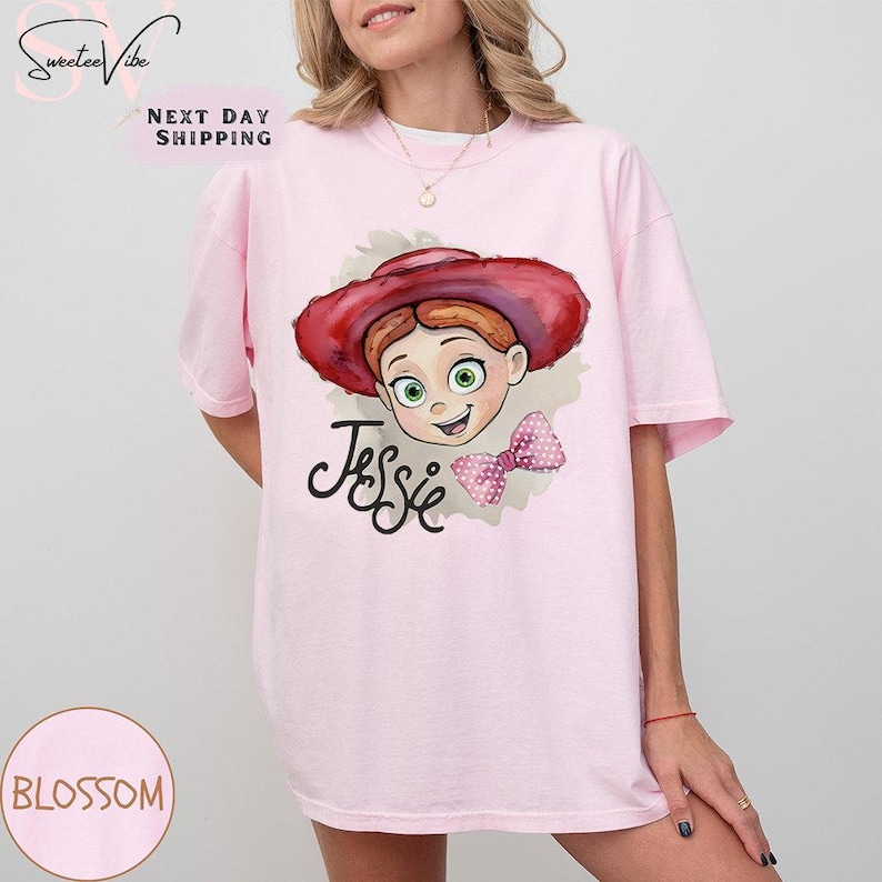 Puede incluir: Camiseta rosa claro con una ilustraci&oacute;n de dibujos animados de Jessie de Toy Story. El dise&ntilde;o incluye la cara de Jessie, un sombrero rojo, un lazo rosa y el nombre "Jessie" en escritura negra. La camiseta tiene mangas cortas.