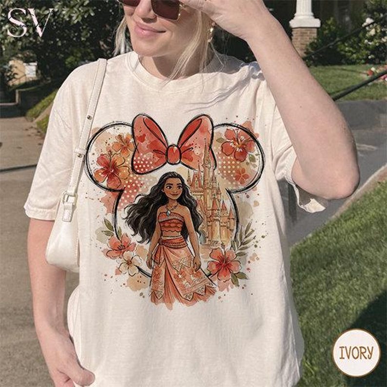 May include: Ivory-colored t-shirt featuring a Minnie Mouse-inspired design with a large bow and floral accents. The graphic includes a Moana character and a castle, creating a whimsical and colorful design.