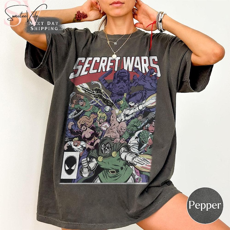 Puede incluir: Camiseta gris oscuro con una ilustraci&oacute;n de c&oacute;mic que dice "SECRET WARS". El gr&aacute;fico incluye varios personajes en colores vibrantes, con una pancarta roja y blanca en la parte superior. Se ve un logo de Spider-Man.