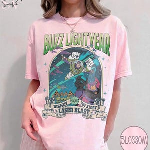 Puede incluir: Camiseta rosa claro con un gr&aacute;fico de Buzz Lightyear de Toy Story. El dise&ntilde;o incluye el texto "Buzz Lightyear" en verde y "Disney Laser Blast Toy Story" en una pancarta. La camiseta tiene mangas cortas.