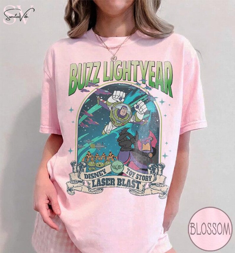 Puede incluir: Camiseta rosa claro con un gr&aacute;fico de Buzz Lightyear de Toy Story. El dise&ntilde;o incluye el texto "Buzz Lightyear" en verde y "Disney Laser Blast Toy Story" en una pancarta. La camiseta tiene mangas cortas.