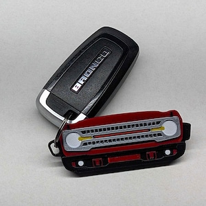 May include: A red and black keychain shaped like a car's front grill, with white and yellow accents. The keychain is attached to a black car key with the word "BRONCO" on it.
