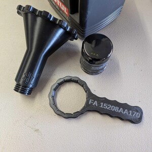 Puede incluir: Herramientas automotrices negras que incluyen un embudo, un filtro de aceite y una llave. La llave est&aacute; etiquetada con "FA 15208AA170". El filtro de aceite es negro con texto plateado. El embudo tiene un borde acanalado y una base roscada.