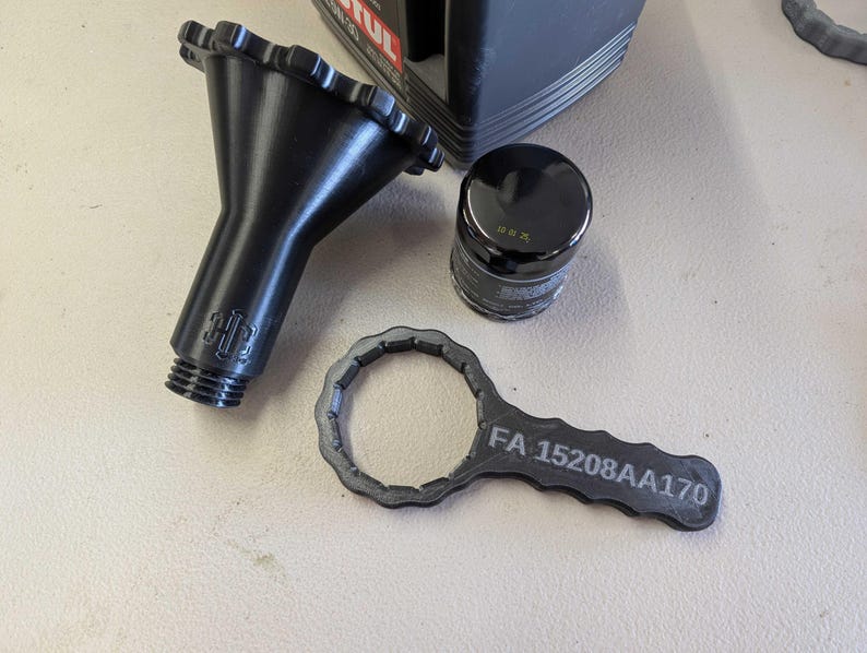 Puede incluir: Herramientas automotrices negras que incluyen un embudo, un filtro de aceite y una llave. La llave est&aacute; etiquetada con "FA 15208AA170". El filtro de aceite es negro con texto plateado. El embudo tiene un borde acanalado y una base roscada.