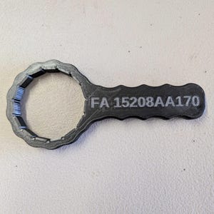 Puede incluir: Una llave de color gris oscuro con una cabeza circular y un mango. El texto "FA 15208AA170" est&aacute; impreso en el mango. La llave est&aacute; sobre una superficie de color claro.