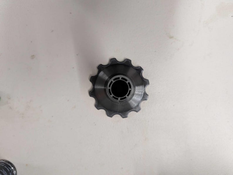 Puede incluir: Una polea de cambio de bicicleta de color gris oscuro con un borde exterior dentado y una abertura central circular. La polea est&aacute; hecha de un material met&aacute;lico y est&aacute; dise&ntilde;ada para su uso en el sistema de transmisi&oacute;n de una bicicleta.