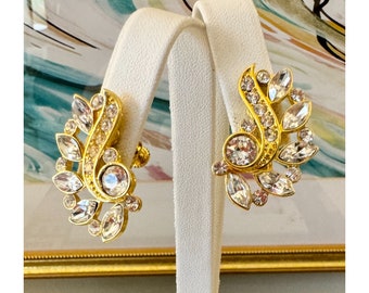 May include: A pair of gold tone clip-on earrings with clear rhinestone accents. The earrings are shaped like a stylized leaf with a cluster of rhinestones.