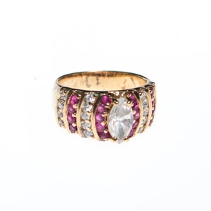 May include: A gold ring with a large, clear, marquise-shaped center stone surrounded by smaller clear stones and red stones.