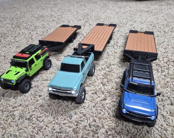 May include: Three toy trucks in lime green, light blue, and dark blue, each pulling a flatbed trailer with a wood-grain textured surface. The lime green truck has a roof rack, the light blue truck has a black hitch, and the dark blue truck is a Bronco.