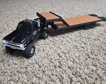 May include: A black pickup truck model with a chrome front bumper and grille, featuring illuminated headlights. The truck is attached to a long, flatbed trailer with a tan wooden-style deck and black frame, equipped with dual rear wheels.