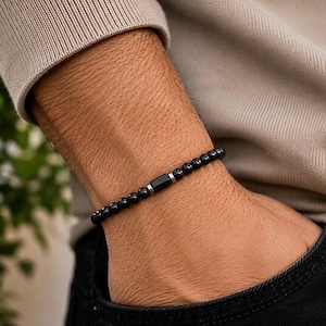 May include: A beaded bracelet featuring small, round, polished black beads and a single, central, rectangular black bead. The beads are separated by tiny, metallic silver-toned spacers, creating a uniform and sleek design around the wrist.