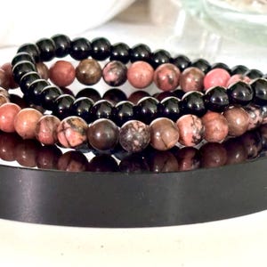 May include: Three beaded bracelets. One bracelet features black beads, while the other two have a mix of pink, brown, and tan beads. The bracelets are stacked on a black, reflective surface, creating a layered effect. The beads appear to be natural stone.