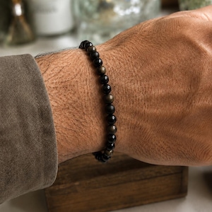 Könnte beinhalten: Ein Perlenarmband bestehend aus abwechselnd glatten, schwarzen, kugelförmigen Perlen und metallisch goldfarbenen Obsidianperlen. Das Armband ist auf einem flexiblen Band aufgezogen, wodurch es eng am Handgelenk anliegt. Die Perlen sind gleichmäßig.