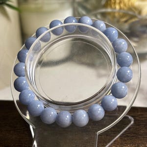 May include: A circular bracelet composed of smooth, round, light blue gemstone beads. The beads exhibit a consistent pale blue hue with subtle internal variations and a polished finish. The bracelet is displayed on a clear, circular stand.