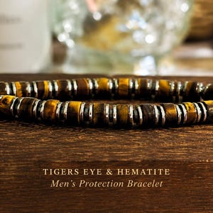 May include: A beaded bracelet featuring alternating cylindrical tiger's eye stones and thin metallic hematite spacers. The text TIGERS EYE & HEMATITE Protection Bracelet is printed below the jewellery piece on the dark wooden surface.